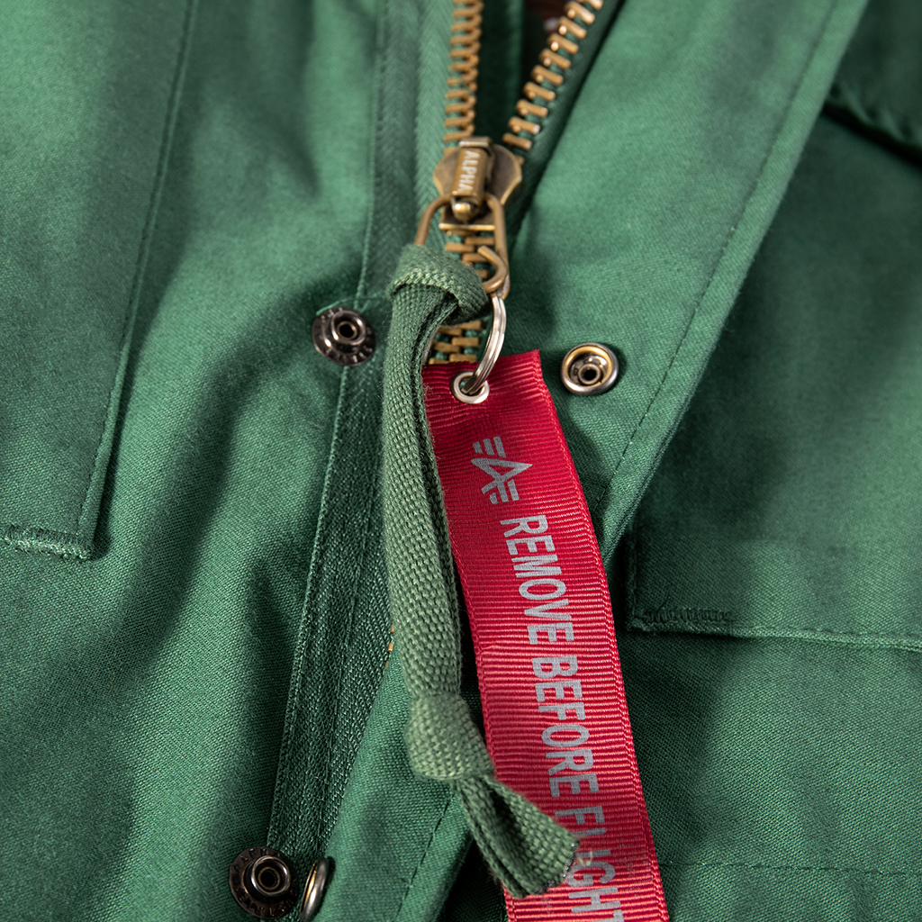 Detail of the heavy-duty brass zipper and "Remove Before Flight" tag on the custom olive green M65 field jacket.
